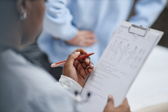 Black woman holding clipboard reviewing medical chart with anatomical diagrams while standing near Caucasian woman in clinical setting, focusing on healthcare documentation and patient assessment