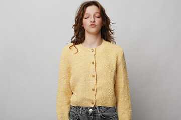 Young woman in yellow knitted cardigan stands with closed eyes and relaxed expression against grey...