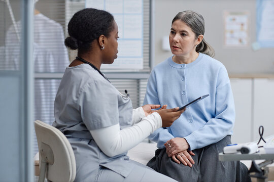 Black female nurse discussing medical information with middle aged Caucasian woman in clinical setting, nurse holding digital tablet while patient listening attentively during consultation