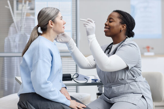 Black female young adult healthcare worker examining eyes of Caucasian middle aged woman using medical penlight during checkup in modern clinic setting