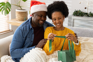 African American couple sitting on sofa at home, sharing green gift box and chunky knit blanket