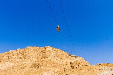 Obraz premium Cable car ascending to Masada fortress in Judean Desert, Israel. Ancient historic site near Dead Sea under bright blue sky.