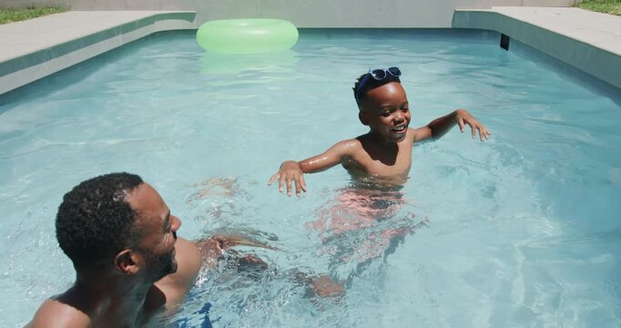 Mature male guardian splashing arm, guiding child practicing kicks in backyard pool with green ring
