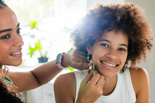 African American women fastening earring while seated woman holding green gua sha at window