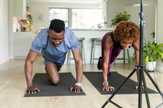 Diverse couple holding planks on mats at home by tripod wearing fitness watches in sportswear