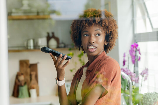 African American woman in rust shirt holding device to mouth, wearing smartwatch in bright kitchen