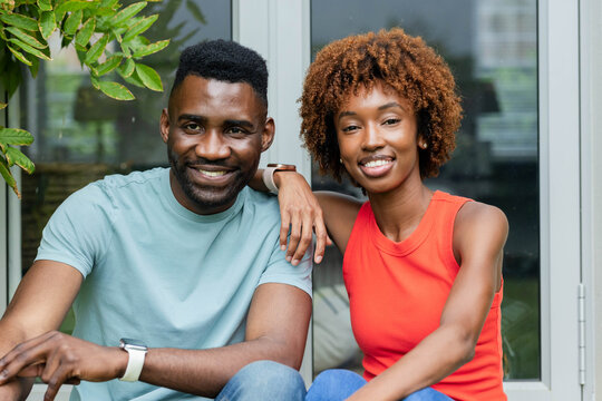 Diverse couple sitting on house threshold wearing casual clothes with smartwatch and plant