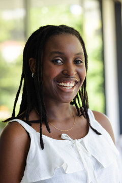 African woman smiling, standing near large windows, wearing white ruffled blouse, gold necklace