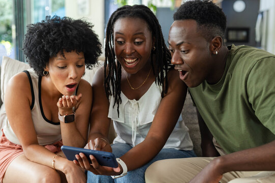 African American friends leaning forward, gasping at blue-cased smartphone on light sofa