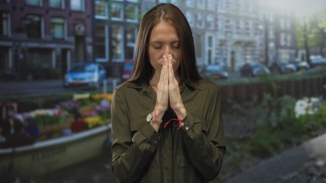 Woman with hands pressed together in prayer, closed eyes, red string bracelet and wristwatch, standing on an amsterdam street by a canal; serenity.