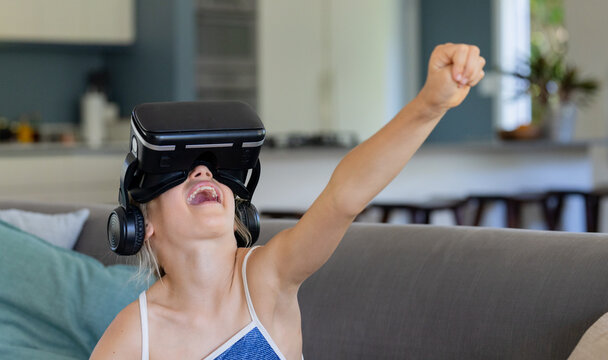 Youth female sitting on gray couch at home wearing black VR headset and headphones, laughing