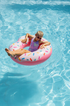 Female child reclining on pink donut float, wearing one-piece swimsuit and pink sunglasses in pool