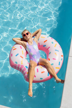 Female youth reclining on pink donut float wearing lilac swimsuit and red shades near pool coping