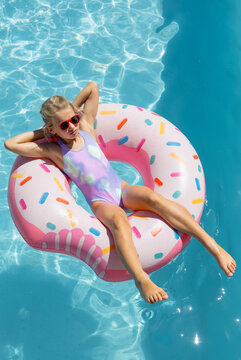 Child reclining on pink sprinkle donut float in blue pool, wearing red sunglasses and swimsuit