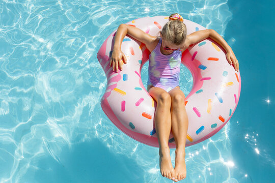 Child girl floating in pool on pink donut float with sprinkles, in pastel swimsuit and sunglasses