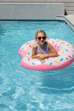 Child girl floating on pink donut float in pool wearing red sunglasses and lavender swimsuit