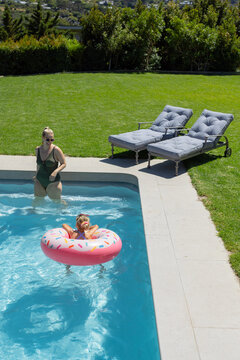 Adult woman in green swimsuit standing in backyard pool while child floating on pink donut