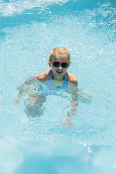 Female child wading in outdoor pool water wearing purple swimsuit and red round sunglasses