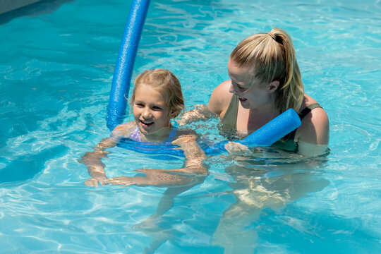Female adult supporting female child in outdoor pool, holding blue foam noodles, wearing swimsuits