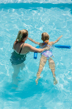 Woman supporting child girl floating on blue pool noodle in outdoor pool, dark swimsuit, scrunchie