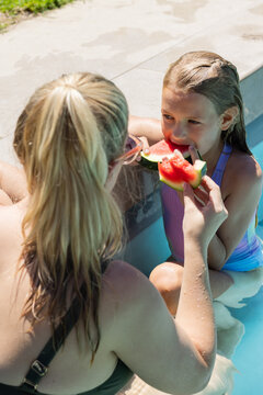 Female caregiver and child sharing large watermelon slice at backyard pool edge in swimsuits