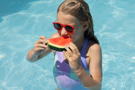 Child female standing waist-deep in pool, biting watermelon slice in lilac swimsuit and red shades