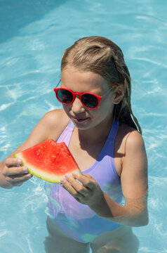 Preteen girl holding watermelon slice while standing in pool in purple swimsuit and red sunglasses