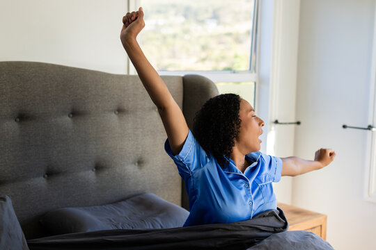 Female sitting up stretching and yawning in bedroom wearing blue pajama top with gray duvet
