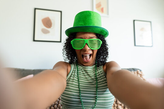 African American woman taking low-angle selfie on sofa wearing green glitter hat, shades and beads