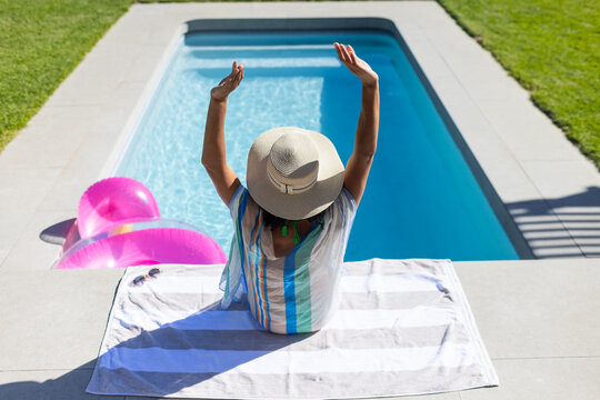 Adult woman of African descent sitting on striped towel at pool edge, straw hat, pink rings