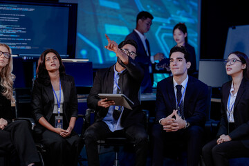 Obraz premium A large, diverse team of business professionals in a group meeting. An Indian man with a tablet leads the discussion with Asian and Caucasian colleagues in a high-tech office.