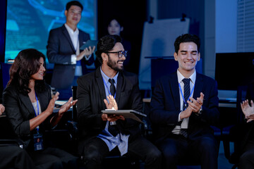 Obraz premium A large, diverse team of business professionals in a group meeting. An Indian man with a tablet leads the discussion with Asian and Caucasian colleagues in a high-tech office.