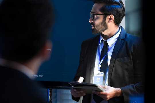 A tech expert, an Indian man, presents a new AI-driven software solution at a business summit. He holds a tablet and engages with the audience during an interactive QA session.