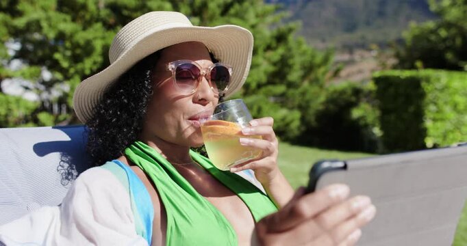 African American woman in sunhat reclining on lawn chair, tapping tablet, sipping glass with orange