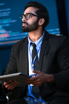 An Indian male software engineer or data scientist holds a tablet in a command center. He is asking a question about the AI code or cybersecurity data shown on the large screen.
