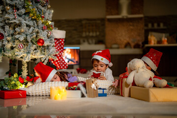 Three kids in Santa hats gather under a Christmas tree, surrounded by glowing presents and cozy decorations. They open gift boxes and share festive joy in a warm holiday setting.