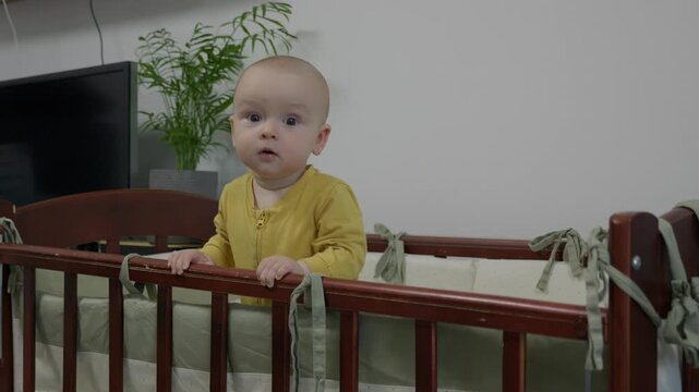 caucasian baby standing at crib smiling broadly, milestone achiever in yellow onesie showing joyful grin and playful gaze, bright nursery with potted plant and modern decor capturing