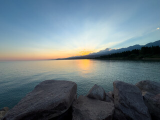 Obraz premium Scenic sunset over Issyk Kul Lake in Kyrgyzstan. The sun sets behind the mountains, casting a golden reflection across the water.