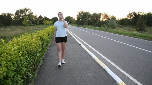 Young woman walking toward camera along sidewalk while drinking water and wearing wireless earbuds at sunset. Dynamic front tracking shot with natural motion creates modern urban fitness atmosphere.
