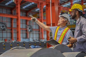 Two factory workers discussing metal coil inspection in an industrial warehouse. They wear safety gear, including helmets and vests, emphasizing teamwork, manufacturing, and industrial safety.
