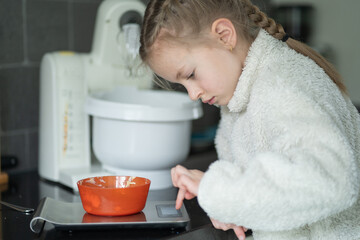 Young girl in white fleece jacket measures ingredients in an orange bowl on a digital kitchen scale, with a stand mixer and kitchen appliances visible in the background © Ann