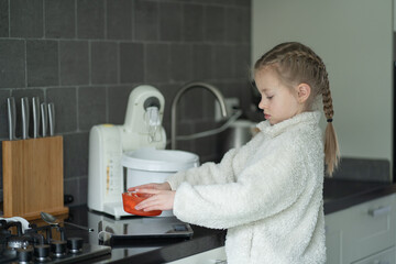 Young girl with braided hair in white fleece sweater prepares ingredients in a modern kitchen, featuring a mixer and gas stove with utensils on a countertop © Ann