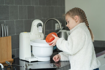 Young girl with braided hair in white fleece sweater adds ingredients to a mixing bowl using an electric mixer in a modern kitchen with gray tiled walls and wooden utensils © Ann