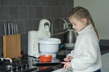 Young girl with braided hair in white fleece jacket measures ingredients in orange bowl on kitchen counter with mixer and utensils visible in modern home kitchen setting © Ann