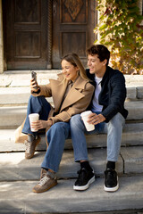 Couple enjoying coffee and taking a selfie on steps outside a building in autumn