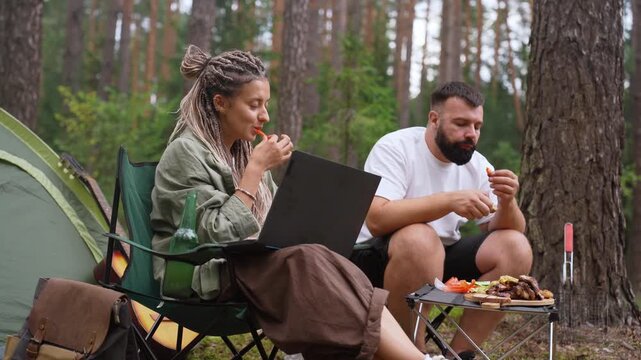 Digital nomadic couple enjoying a meal while working on a laptop at their campsite, surrounded by a tranquil pine forest, combining adventure and outdoor recreation with remote work and a modern lifes
