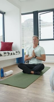 Vertical video: Mature man meditating on green mat in bedroom after prayer pose, centering breath