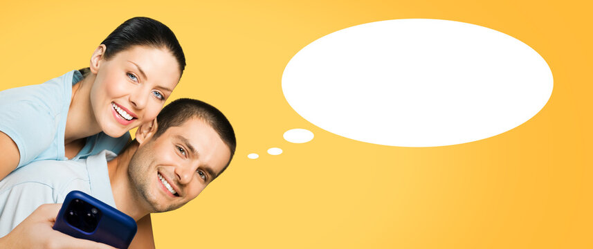 Ad image - expressive smiling couple, man, woman in blue clothing hold cellphone smartphone mobile cell phone, isolated yellow background. White empty think speech bubble, cloud ballon, thought float