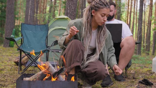Young woman with dreadlocks kindling a fire in a brazier while her freelancer boyfriend works on a laptop at their campsite in a pine forest, enjoying an alternative lifestyle