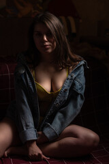 Fototapeta premium Young woman with long dark hair wearing a yellow bra and denim jacket poses in low light, showcasing her features and the texture of the clothing